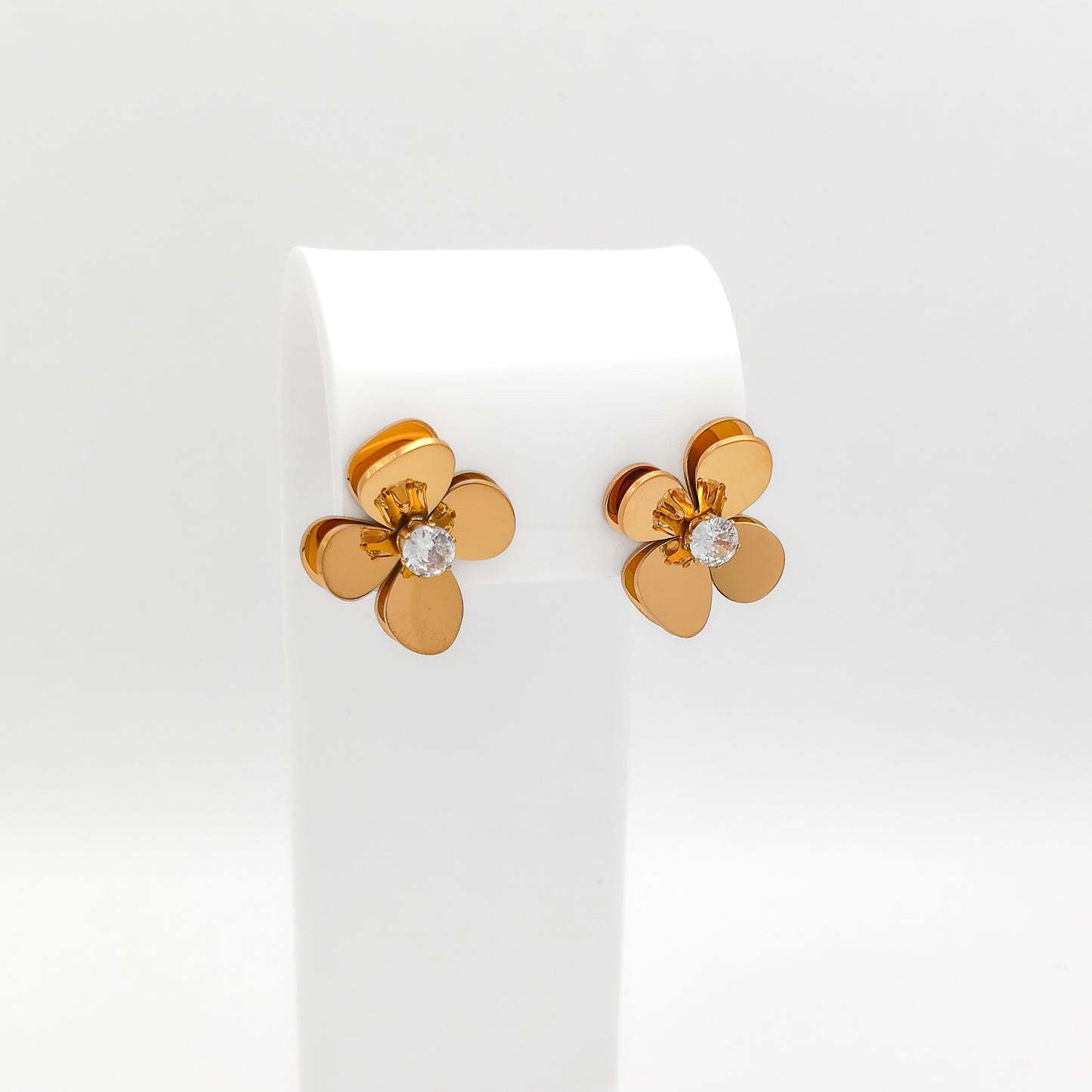 A pair of 18K gold-plated stainless steel 3D floral stud earrings featuring layered, curved heart-shaped petals and a sparkling prong-set round clear cubic zirconia center, displayed on a white stand.