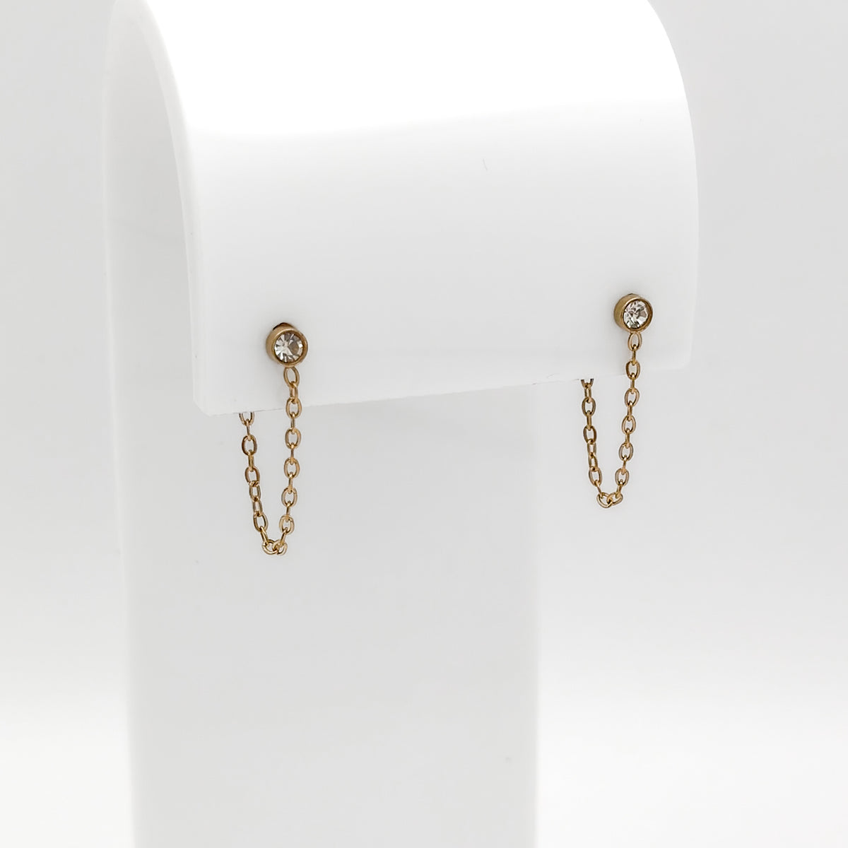 A pair of gold-toned dangle earrings with a small round clear gemstone at the top and a delicate chain hanging below, displayed on a white jewelry stand against a light gray background.