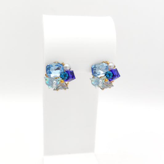 A pair of gold-plated statement stud earrings featuring a cluster of light blue, sapphire, and iridescent crystals, a mini butterfly accent, and a pearl, displayed on a white stand.