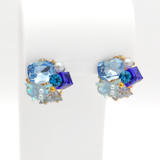 Front view of gold-plated copper alloy cluster stud earrings set with multi-shade blue crystals, pearls, and iridescent accents on a white stand.