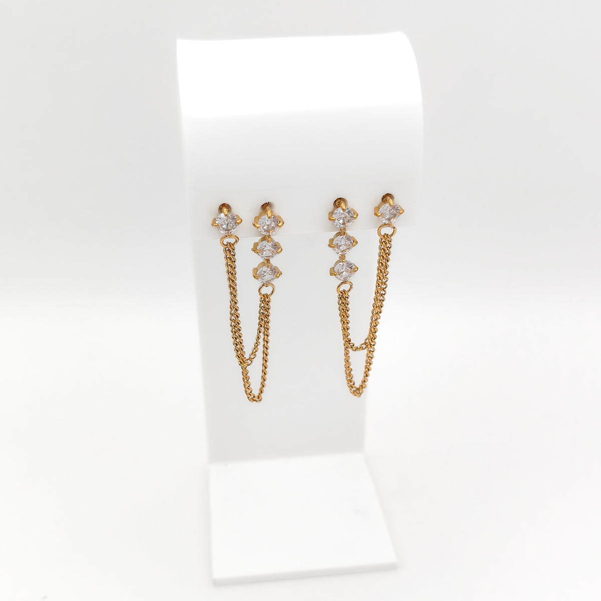 A pair of gold-toned dangle earrings with small clear stones at the top and two hanging chain drops, displayed on a white jewelry stand against a white background.