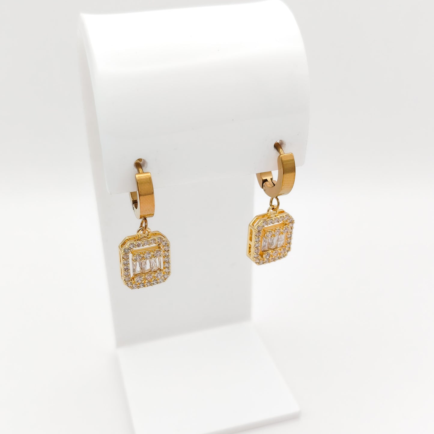 A pair of gold huggie hoop earrings with square drop charms paved with clear stones, displayed on a white jewelry stand against a white background.