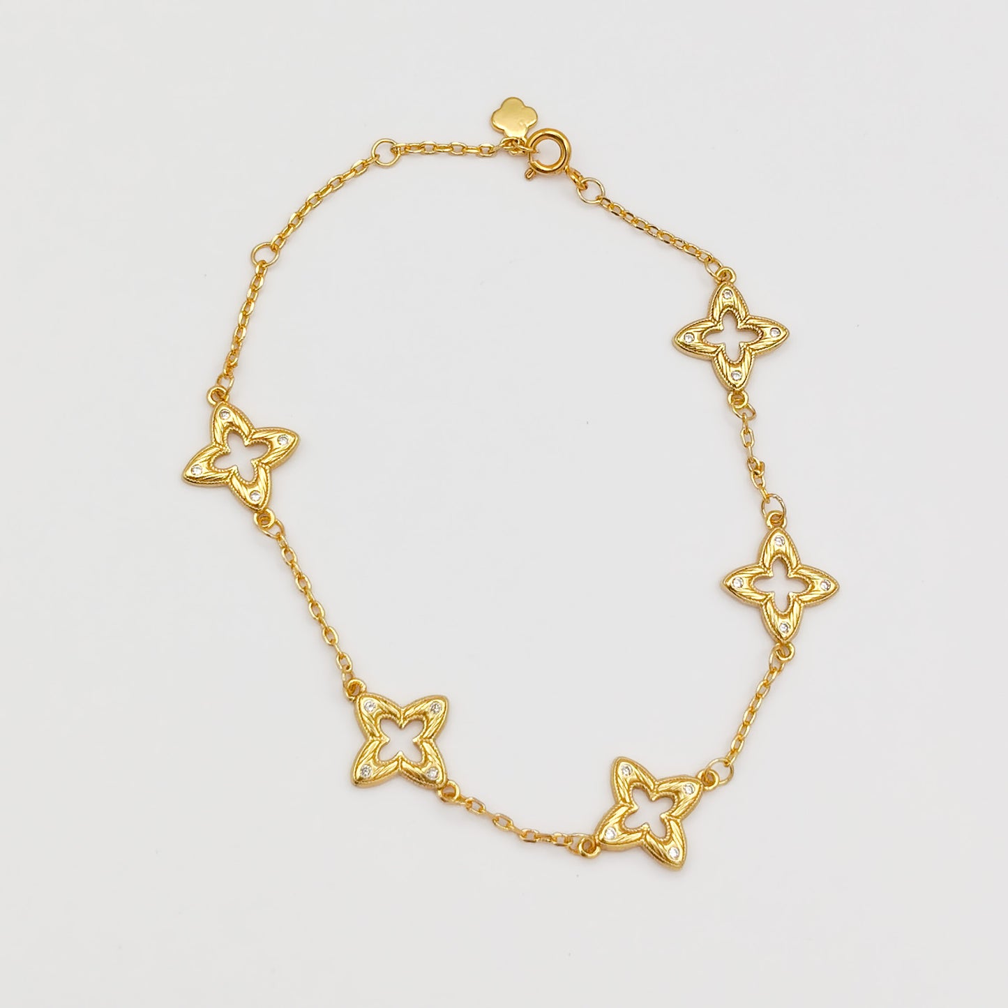 Full view of the gold-plated charm bracelet laid flat, showing five cutout filigree clover links, a spring-ring clasp, and a small flower charm tag.