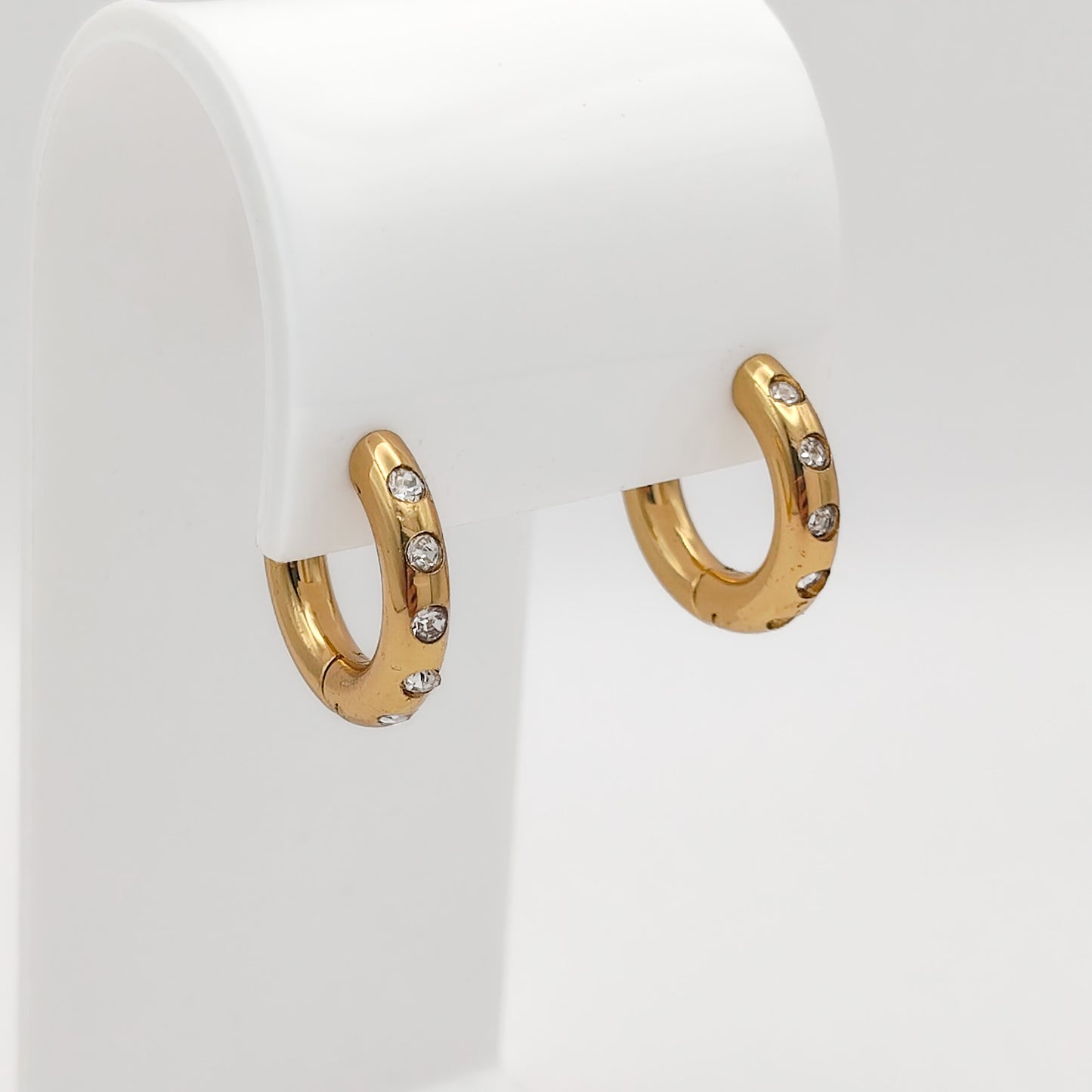 A pair of gold-plated chunky stainless steel hoop earrings inset with glittering clear flush-set rhinestones, displayed on a curved white stand.