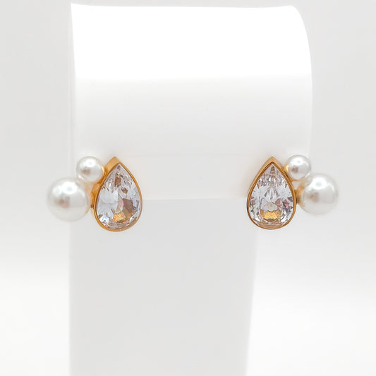 Front view of gold-plated stainless steel stud earrings showcasing a bezel-set pear-cut CZ stone flanked by small white pearls on a white stand.