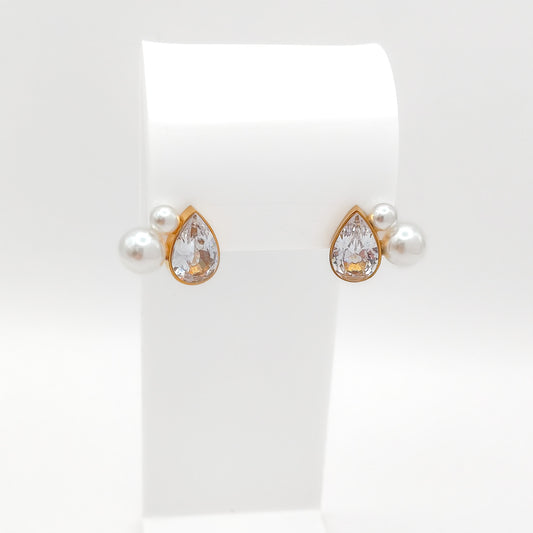 A pair of gold-plated stainless steel cluster stud earrings featuring a bezel-set teardrop cubic zirconia stone accented by two faux pearls, displayed on a white stand.