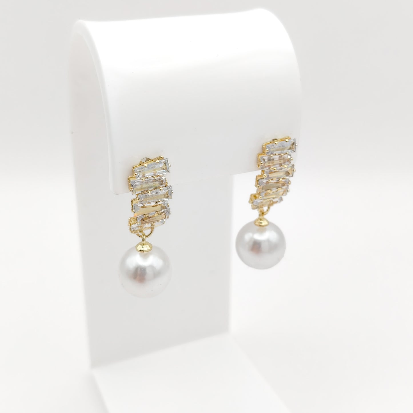 A pair of gold drop earrings with a textured, rhinestone-encrusted top and a single white pearl dangling below, displayed on a white jewelry stand against a white background.