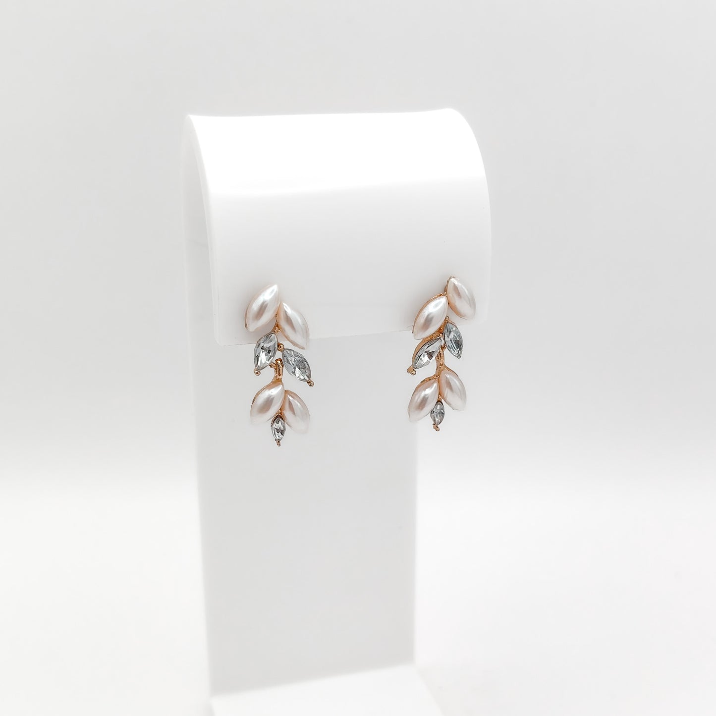 A pair of gold and rose gold leaf drop earrings with white pearls and clear marquise crystals, displayed on a white stand against a light gray background.