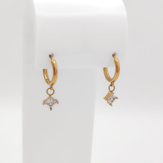 Detailed close-up of a pair of gold-plated stainless steel huggie hoop earrings with dangling square-set princess-cut clear cubic zirconia charms, displayed on a white stand.