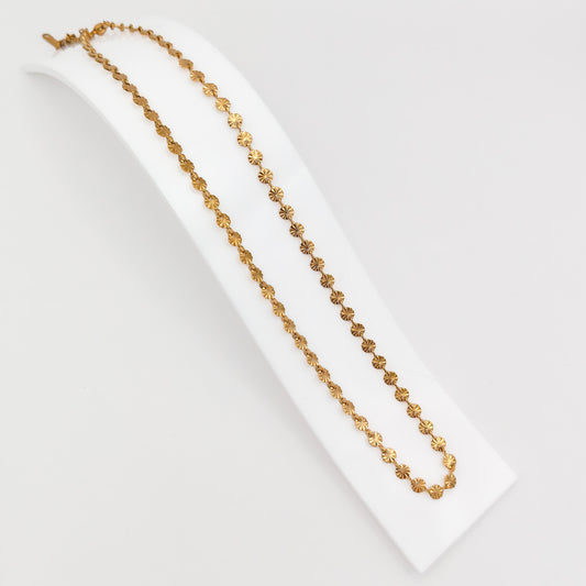 A gold-plated stainless steel princess-length necklace made of a continuous chain of small, textured sunburst disc links, displayed on a curved white stand.