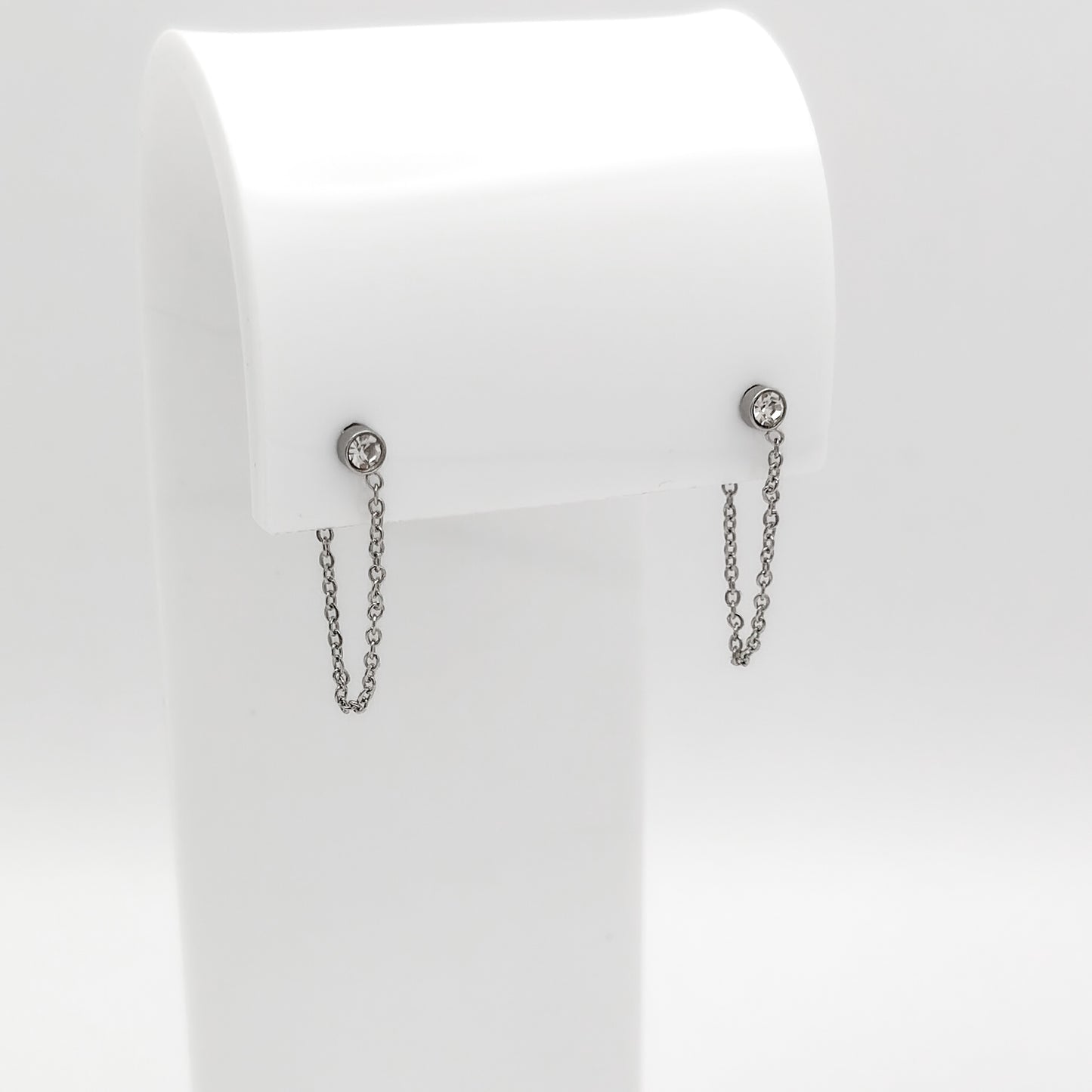 A pair of silver-toned dangle earrings with small round stud tops and short hanging chains, displayed on a white jewelry stand against a light gray background.