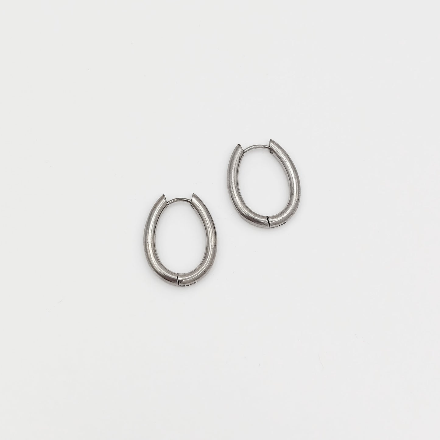 Flat lay photo of a pair of silver-plated stainless steel oval huggie hoop earrings highlighting their smooth, elongated geometric design.