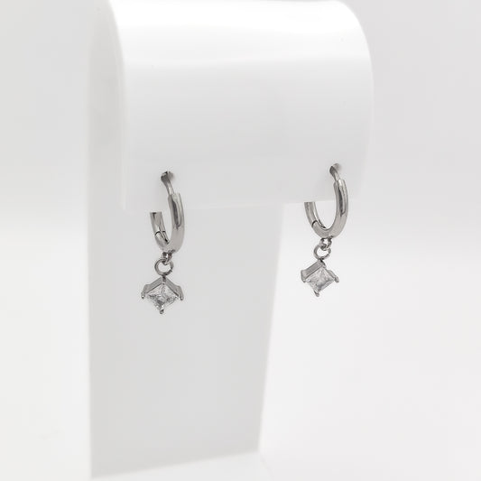 Detailed close-up image of a pair of silver-plated stainless steel huggie hoop earrings featuring dangling square princess-cut clear cubic zirconia charms, displayed on a white stand.