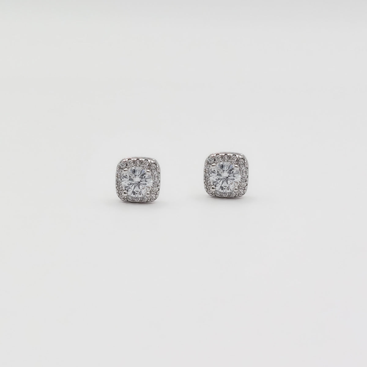Detail image of a pair of square stud earrings with clear gemstones in a silver-toned setting, displayed on a white jewelry stand against a light gray background.