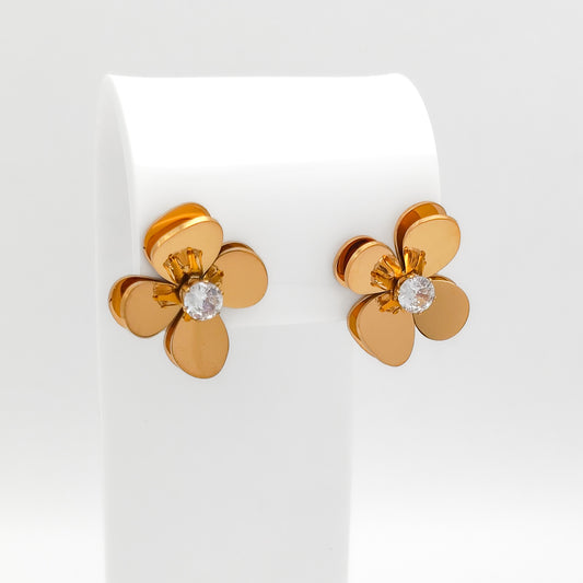 Details of a pair of 18K gold-plated stainless steel 3D floral stud earrings featuring layered, curved heart-shaped petals and a sparkling prong-set round clear cubic zirconia center, displayed on a white stand.
