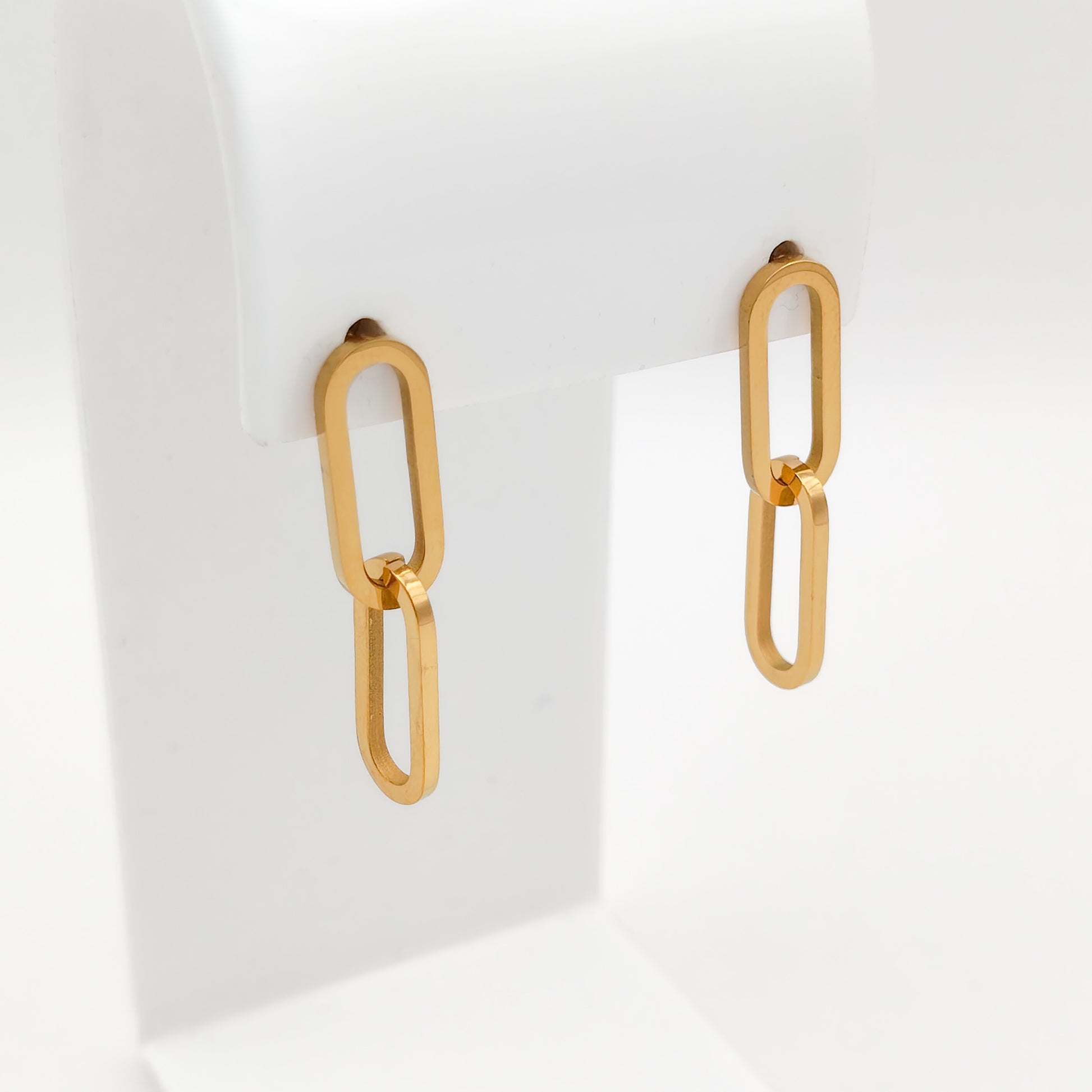 A pair of gold-toned drop earrings with elongated oval links, displayed on a white jewelry stand against a clean white background.