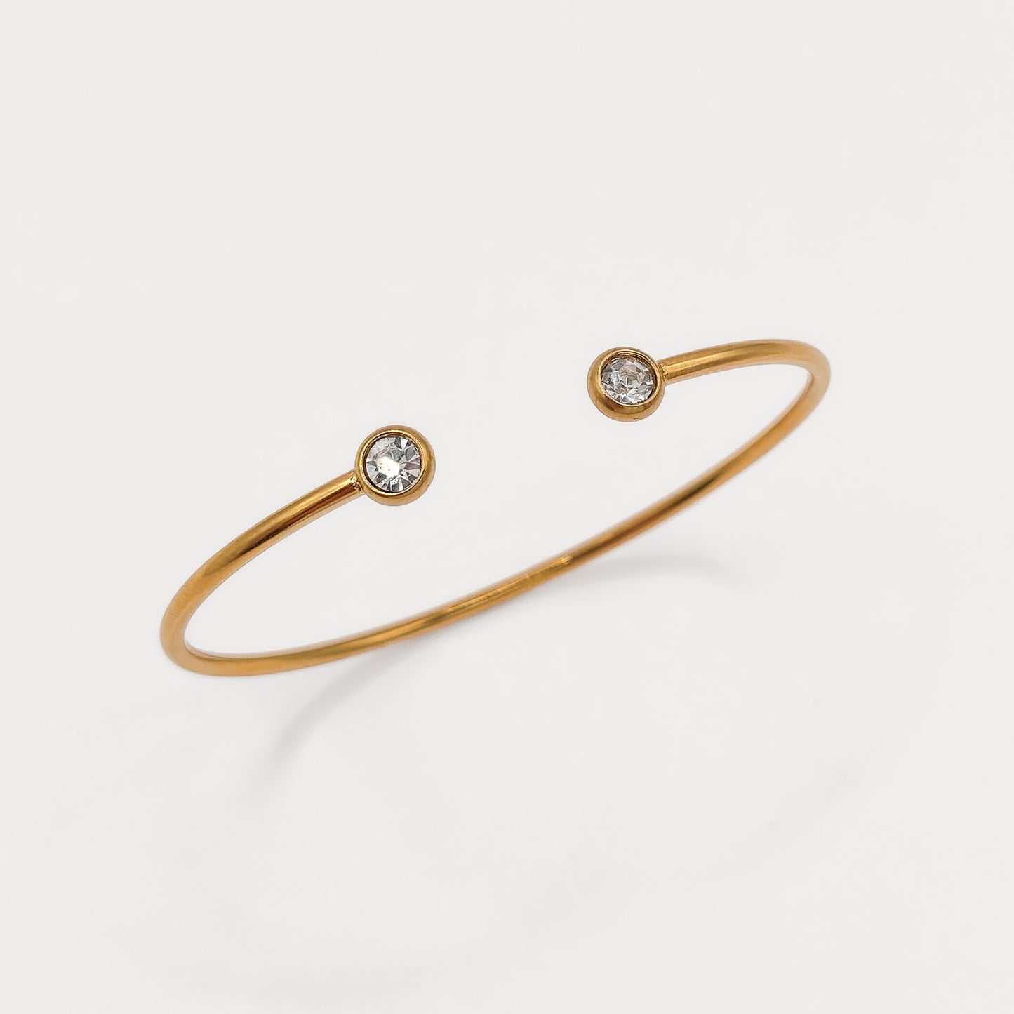A thin gold open cuff bracelet with two small round clear gemstones set at the ends, photographed on a clean white background.