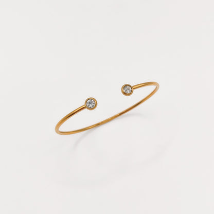 A delicate gold open cuff bracelet with two small bezel-set clear gemstones, displayed on a clean white background.