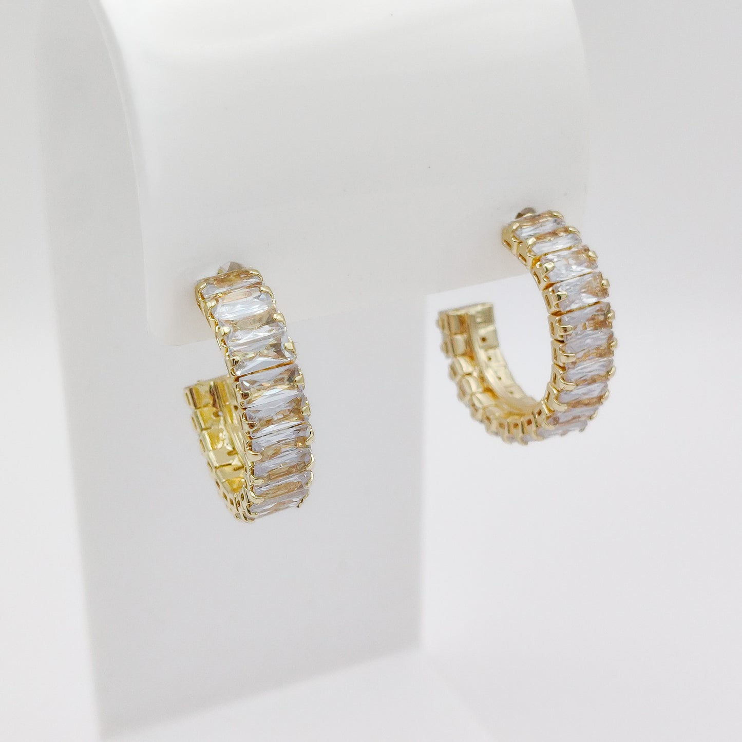 A pair of 25mm gold plated hoop earrings set with clear rectangular baguette crystals, displayed on a white jewelry stand against a white background.