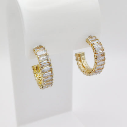 A pair of 25mm gold plated hoop earrings set with clear rectangular baguette crystals, displayed on a white jewelry stand against a white background.