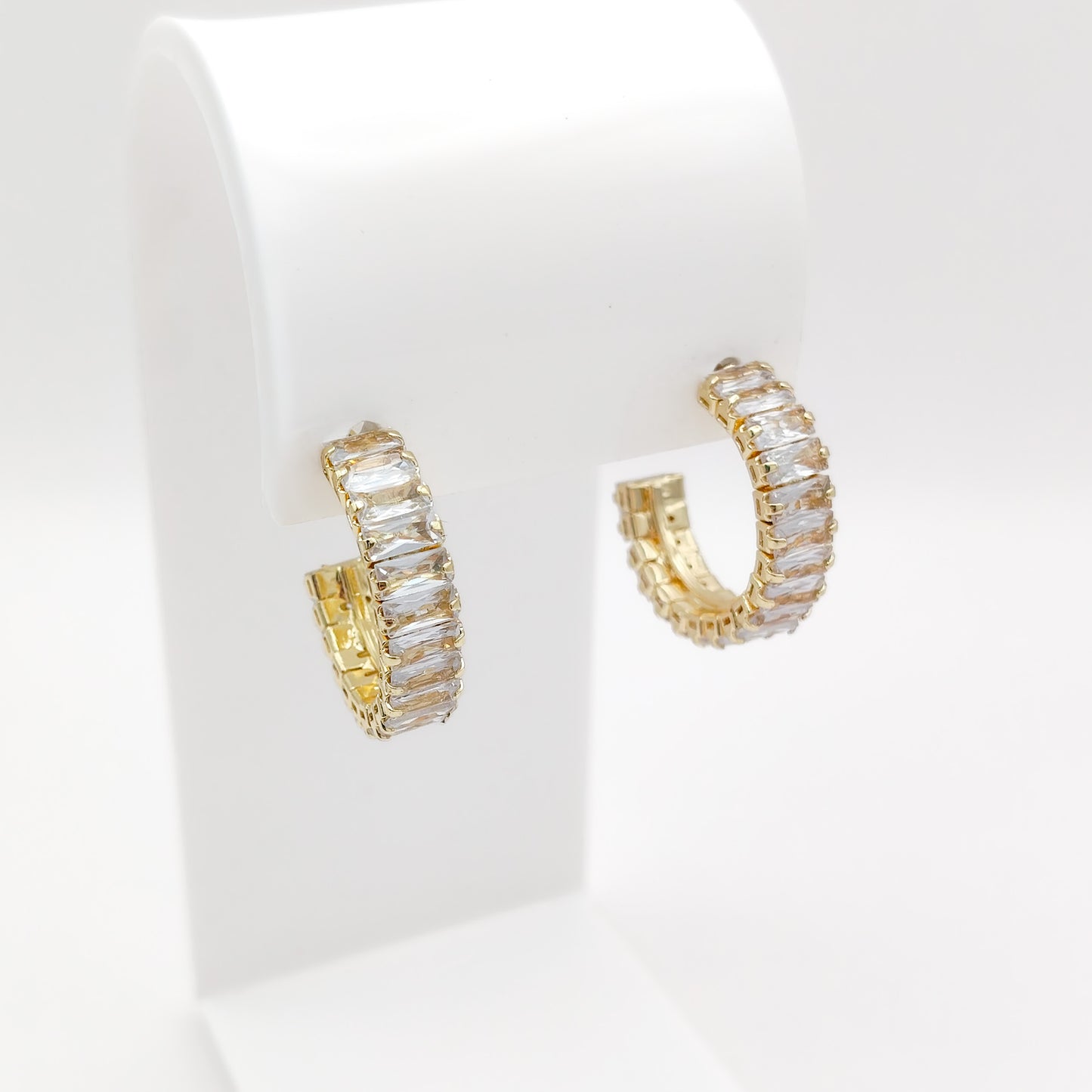 A pair of 25mm gold plated hoop earrings with rectangular clear stones, displayed on a white jewelry stand against a white background.