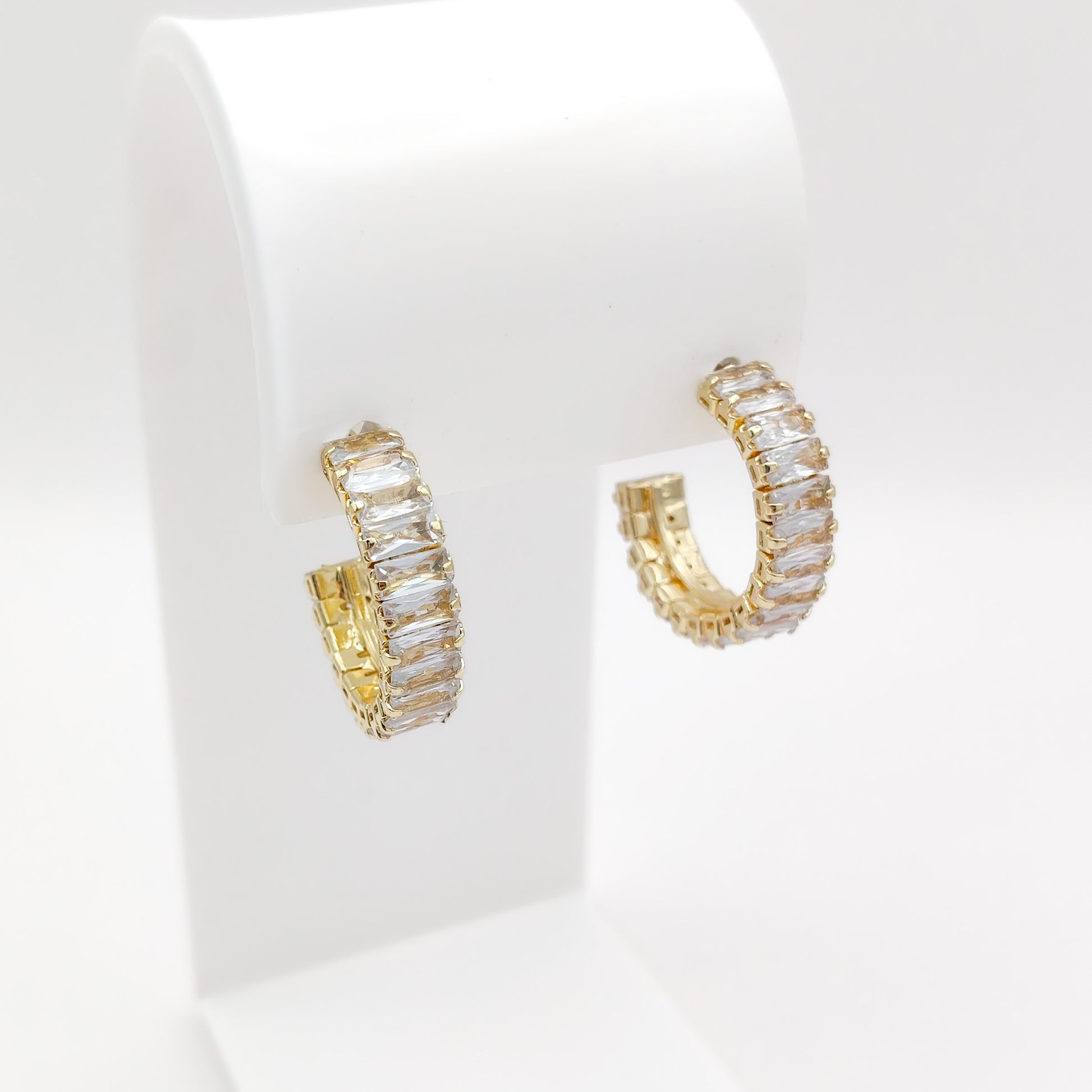 A pair of 25mm gold plated hoop earrings with rectangular clear stones, displayed on a white jewelry stand against a white background.