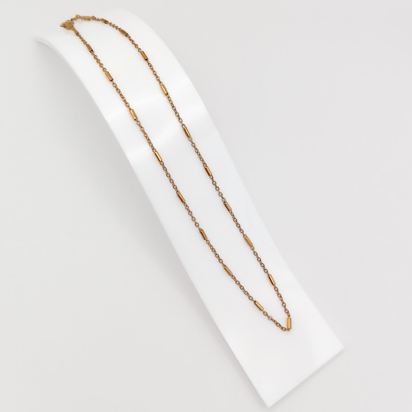 A delicate gold-plated stainless steel chain necklace featuring interspersed cylindrical bar links, displayed on a curved white presentation stand.