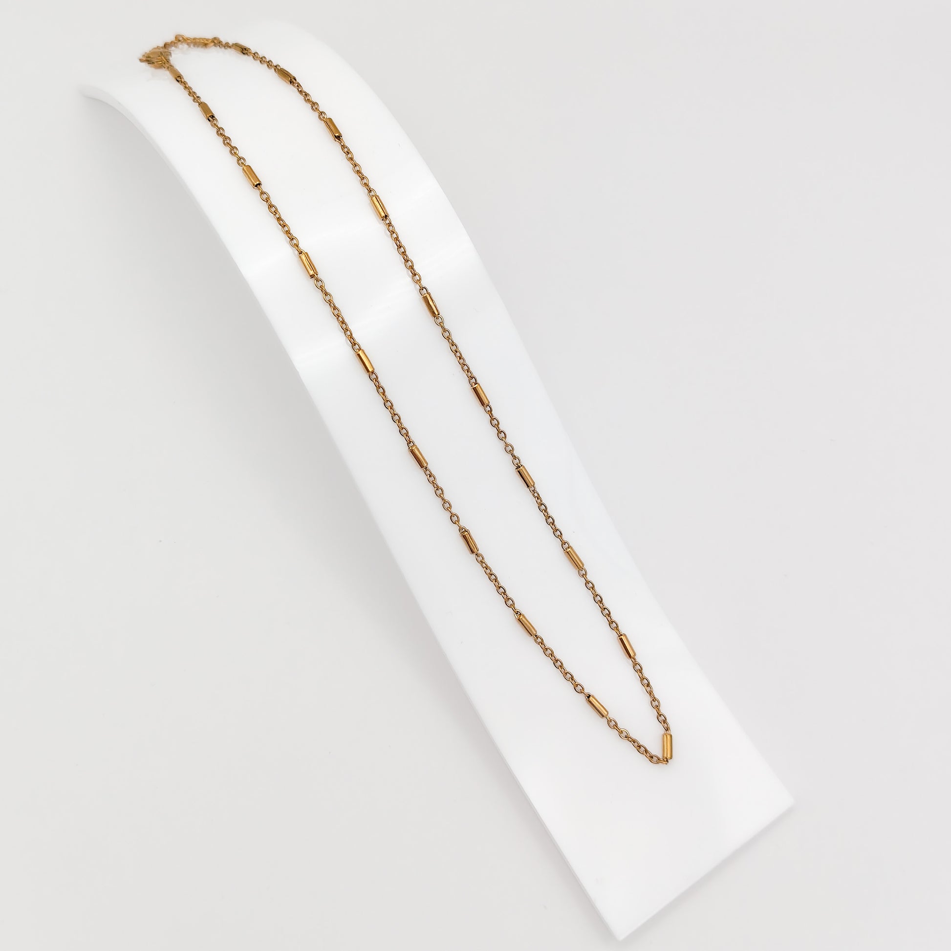 A delicate gold-plated stainless steel chain necklace featuring interspersed cylindrical bar links, displayed on a curved white presentation stand.