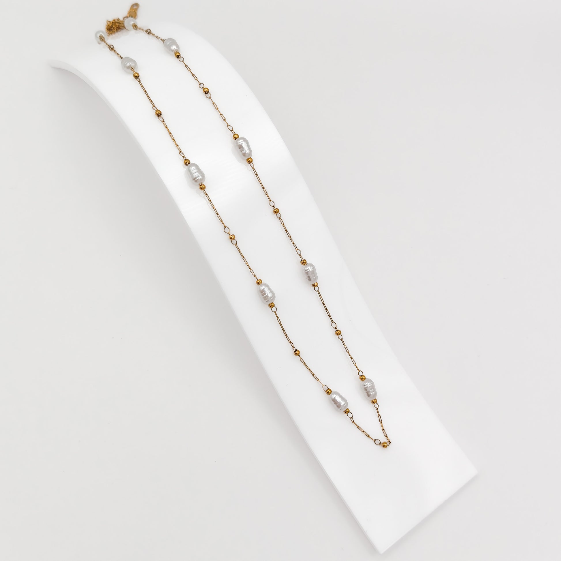 A gold-plated stainless steel princess-length station chain necklace featuring interspersed white baroque imitation pearls and small gold beads, displayed on a curved white display stand.