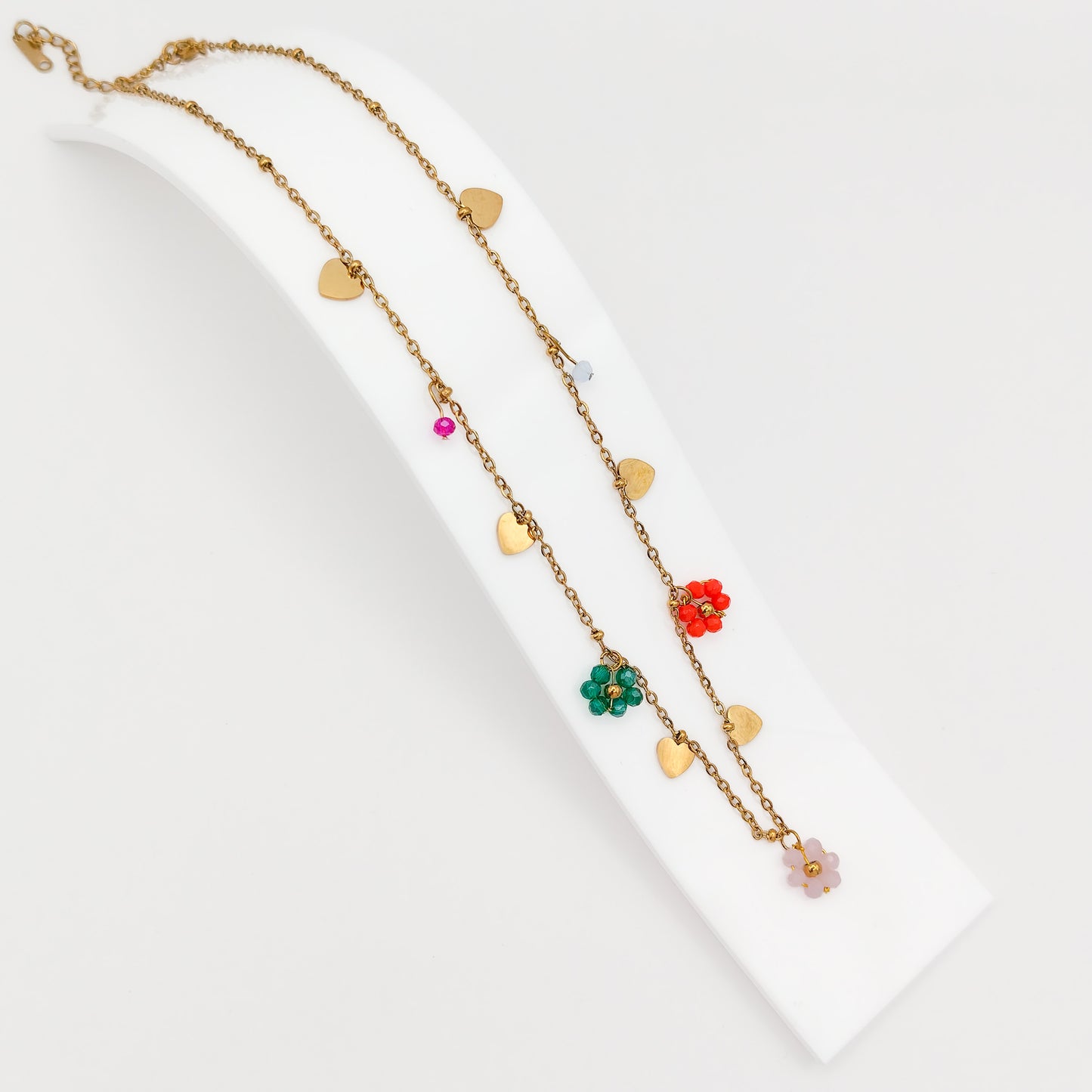 A gold-plated stainless steel princess-length chain necklace adorned with flat gold heart charms and colorful beaded flower charms, displayed on a curved white stand.