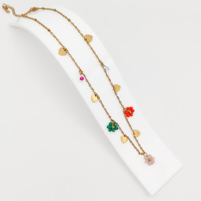 A gold-plated stainless steel princess-length chain necklace adorned with flat gold heart charms and colorful beaded flower charms, displayed on a curved white stand.