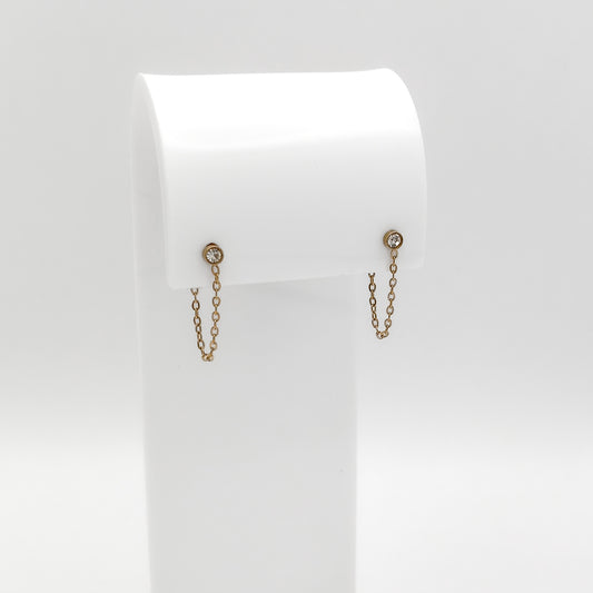 A pair of gold-toned dangle earrings with small circular studs and delicate chain drops, displayed on a white jewelry stand against a light gray background.