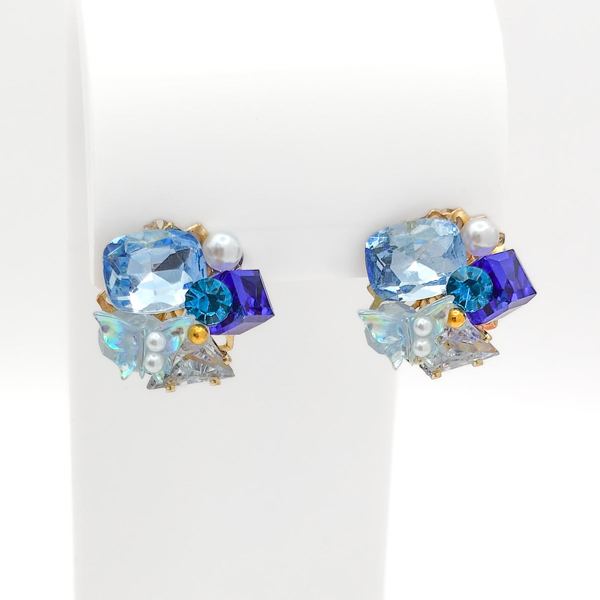 Front view of gold-plated copper alloy cluster stud earrings set with multi-shade blue crystals, pearls, and iridescent accents on a white stand.