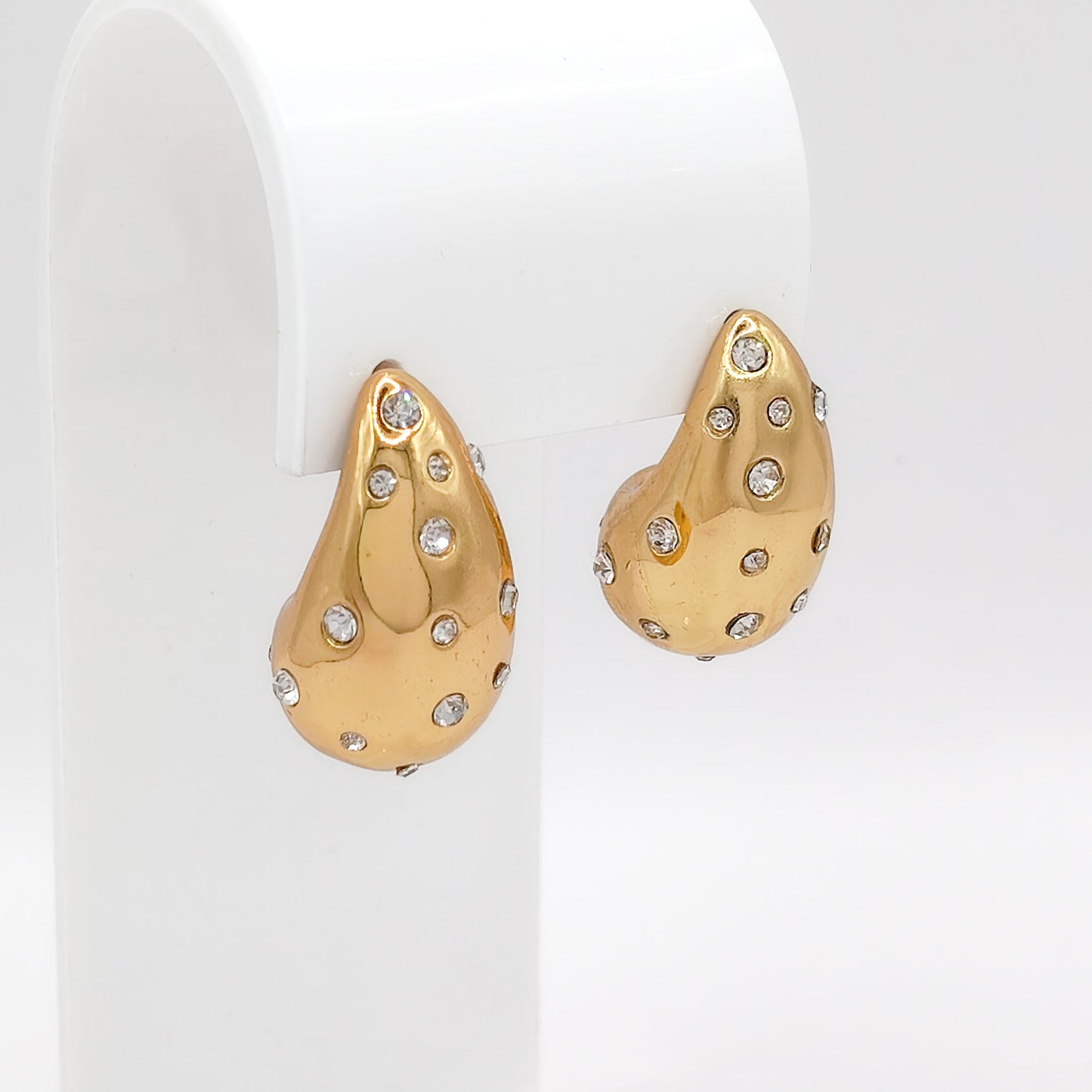 A pair of chunky gold-plated stainless steel teardrop dome stud earrings, scattered with brilliant clear flush-set cubic zirconia stones, displayed on a white stand.
