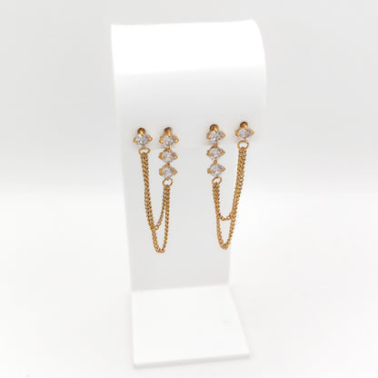 A pair of gold-toned dangle earrings with small clear stones at the top and two hanging chain drops, displayed on a white jewelry stand against a white background.