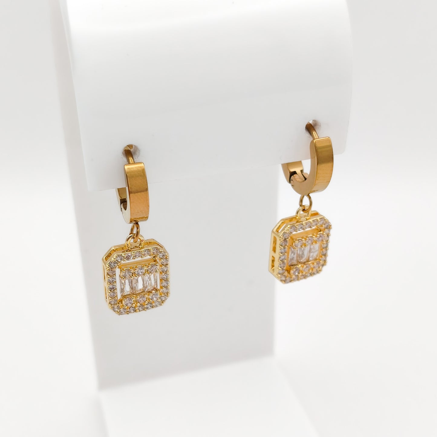 A pair of gold huggie hoop earrings with square baguette-cut clear gemstones, displayed on a white jewelry stand.