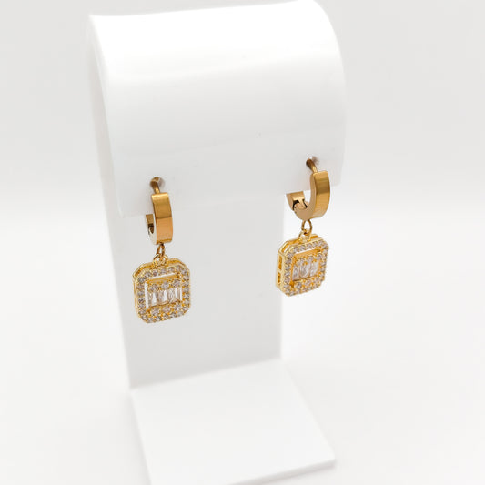 A pair of gold huggie hoop earrings with square drop charms paved with clear stones, displayed on a white jewelry stand against a white background.
