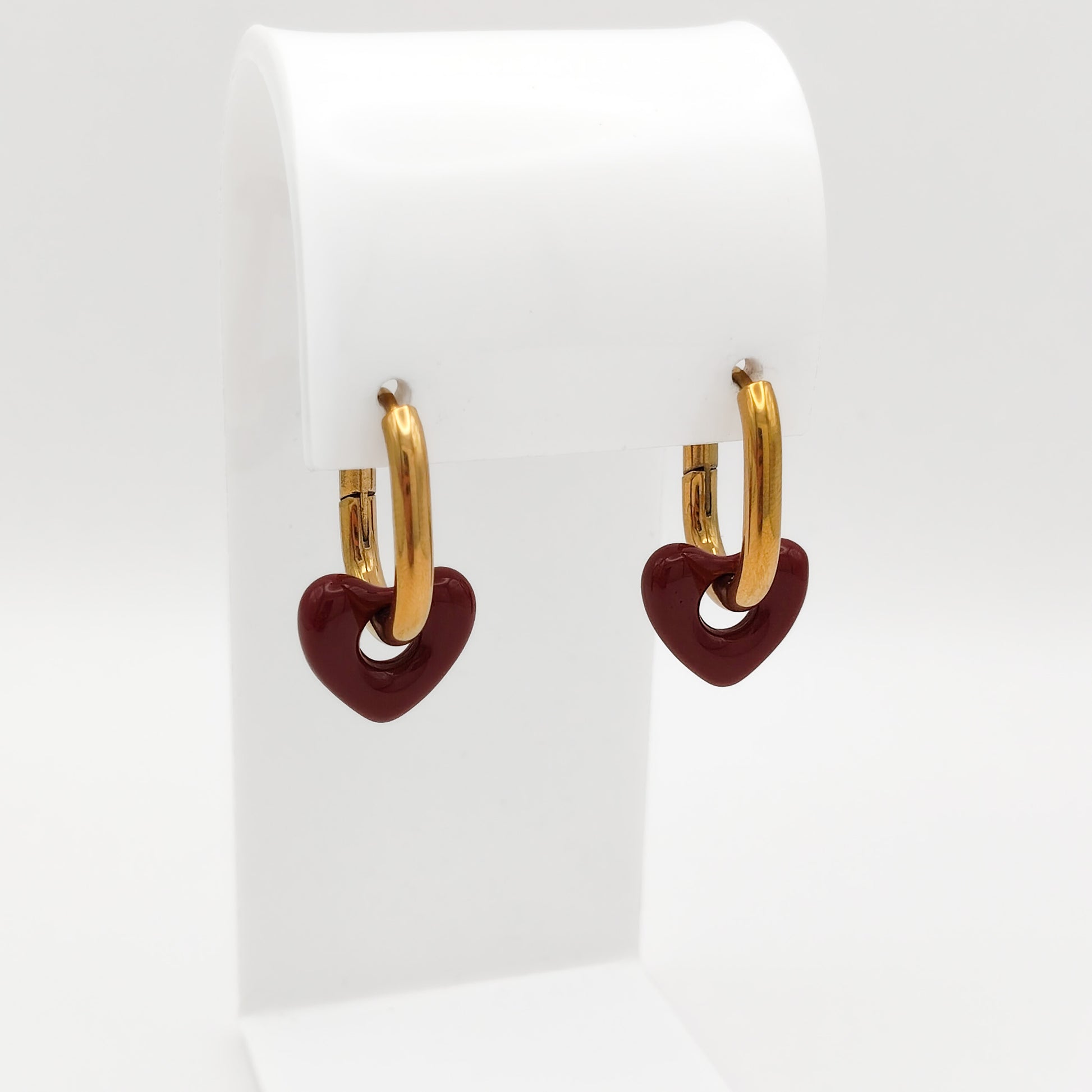 A pair of gold-plated stainless steel huggie hoop earrings featuring dangling burgundy red enamel heart charms, displayed on a white stand.