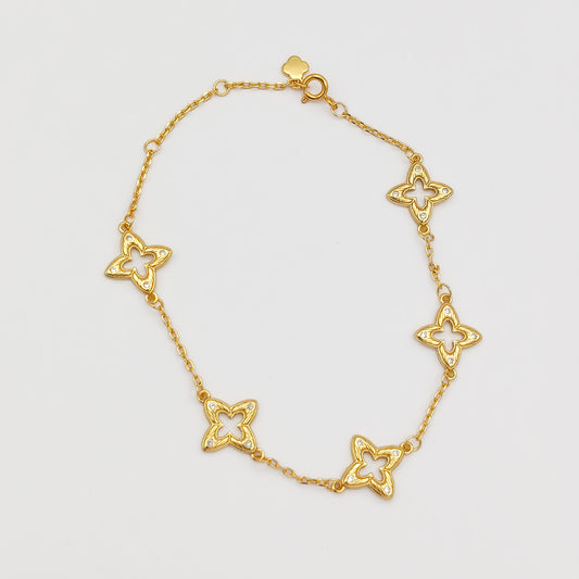 Full view of the gold-plated charm bracelet laid flat, showing five cutout filigree clover links, a spring-ring clasp, and a small flower charm tag.