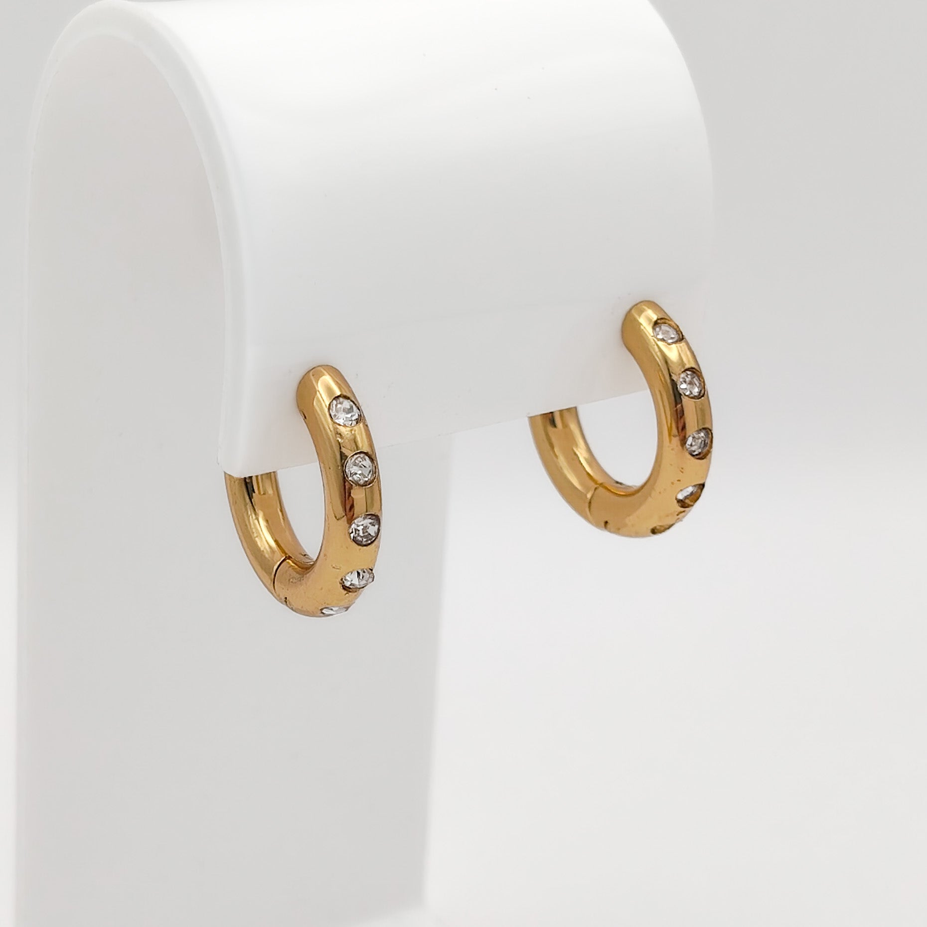 A pair of gold-plated chunky stainless steel hoop earrings inset with glittering clear flush-set rhinestones, displayed on a curved white stand.