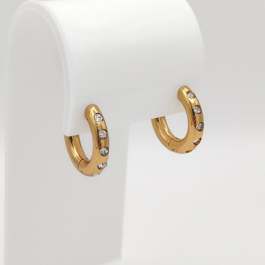 A pair of gold-plated chunky stainless steel hoop earrings inset with glittering clear flush-set rhinestones, displayed on a curved white stand.