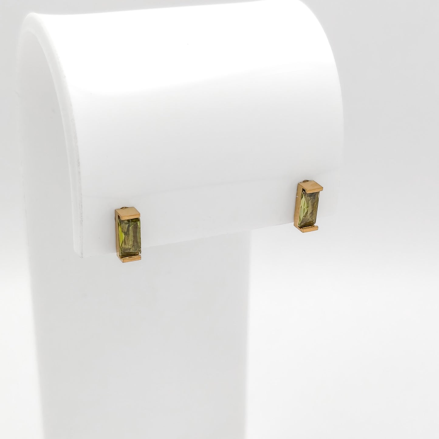 A pair of gold post earrings with rectangular green gemstones, displayed on a white jewelry stand against a white background.