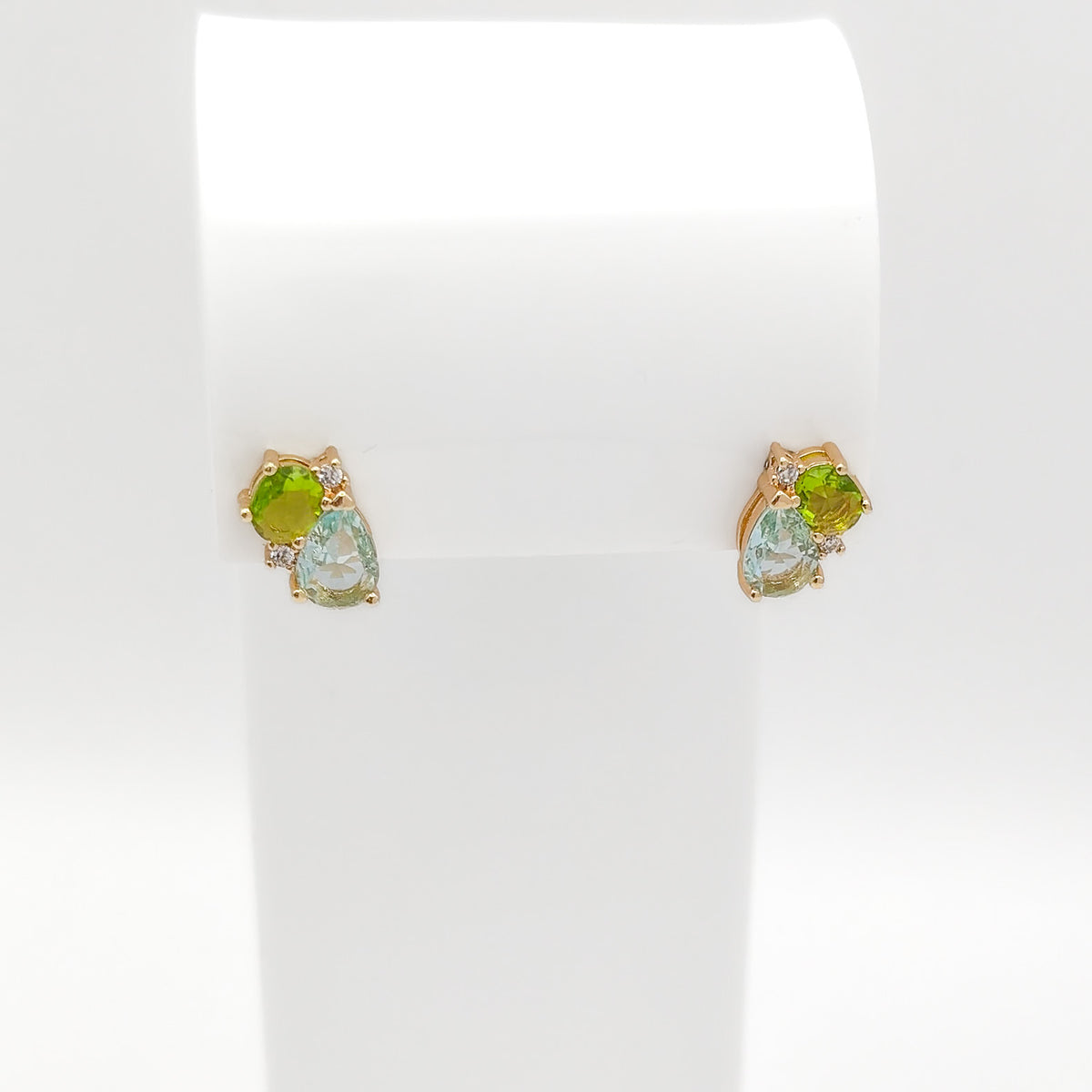 A pair of gold stud earrings displayed on a white jewelry stand. Each earring features a light green marquise-cut gemstone, a round lime-green gemstone, and a small clear accent stone.