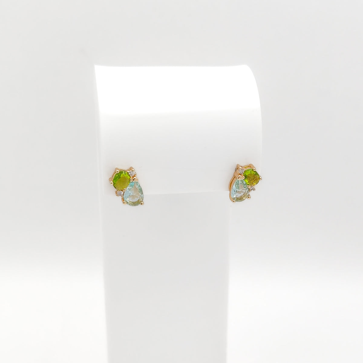 A pair of gold stud earrings with green and iridescent blue-green gemstones, displayed on a white jewelry stand against a white background.
