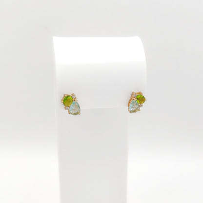 A pair of gold stud earrings with green and iridescent blue-green gemstones, displayed on a white jewelry stand against a white background.