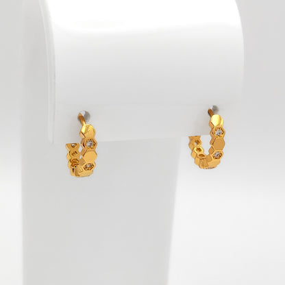 A pair of small gold huggie hoop earrings with a honeycomb textured design and clear crystal accents, displayed on a white jewelry stand.