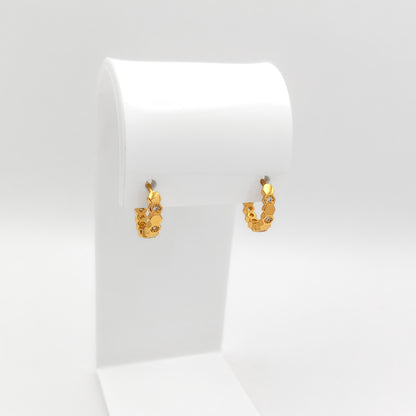 A pair of small gold hoop earrings with tiny clear stones, displayed on a white jewelry stand against a light gray background.
