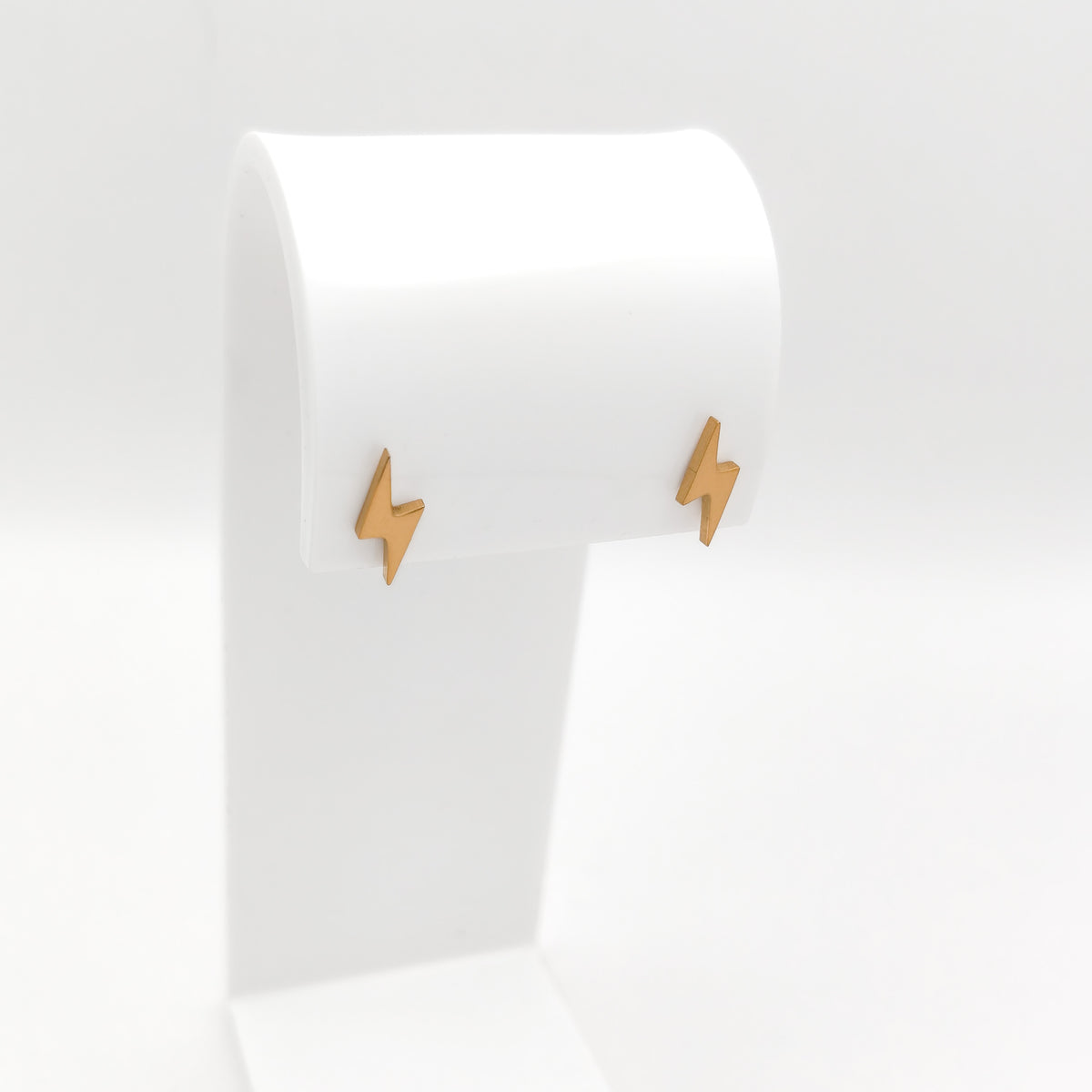 A pair of gold-plated stainless steel lightning bolt stud earrings featuring a polished metallic finish, displayed on a white stand.