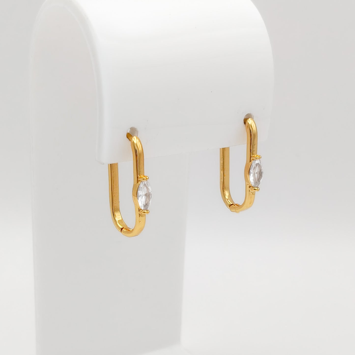A pair of gold plated marquise CZ oval huggie earrings featuring a single bezel-set marquise-cut clear cubic zirconia stone on the front, displayed on a white stand.