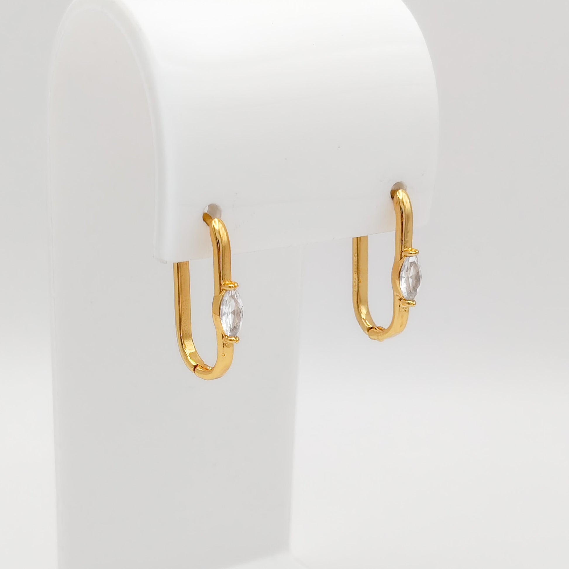 A pair of gold plated marquise CZ oval huggie earrings featuring a single bezel-set marquise-cut clear cubic zirconia stone on the front, displayed on a white stand.